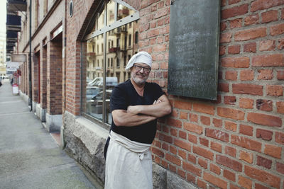 Portrait of senior male baker leaning on brick wall outside bakery