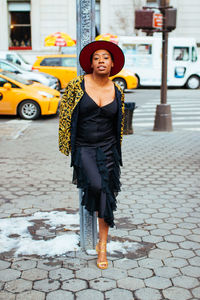 Portrait of woman leaning on pole while standing at footpath in city