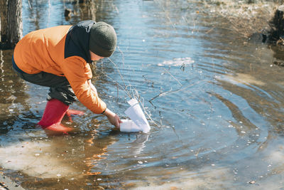 Springtime sailor, amid budding trees, boy leans into experience of launching paper boat, joy