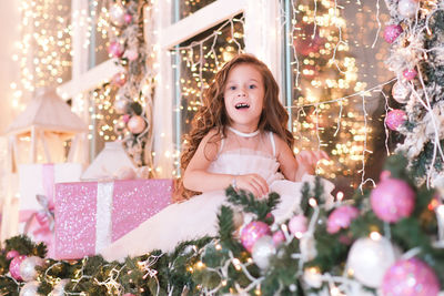 Little beautiful girl sitting on the windowsill, looking and smiling on lights background. new year.