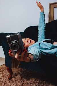 Cheerful woman holding camera lying down on sofa at home