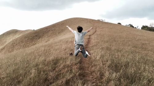 Rear view full length of playful man jumping with arms raised over hill