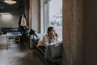 Businesswoman talking over smart phone while looking out through window lying on seat at workplace