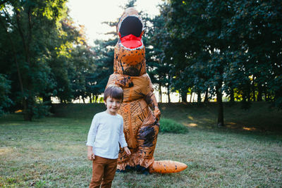 Portrait of boy standing with person wearing dinosaur costume in park