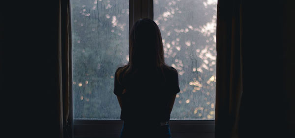 Rear view of silhouette woman looking through window at home