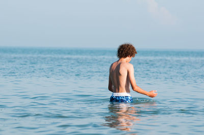 Rear view of shirtless teenage boy standing in sea against clear sky