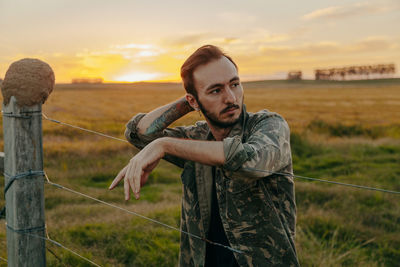 Portrait of young man standing on field against sunset sky
