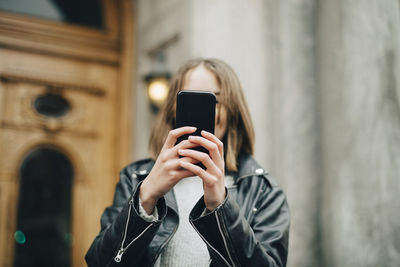 Girl photographing through mobile phone while standing against building