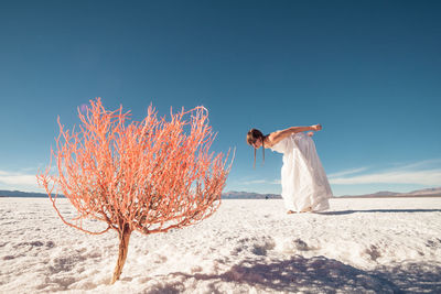 Woman looking at bare tree while bending on snowy field