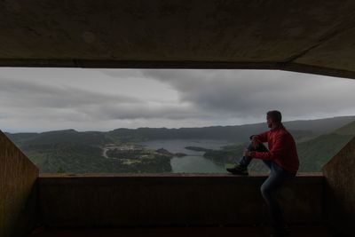 Man looking at view while sitting on retaining wall against sky