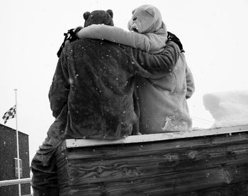 Rear view of friends in costume sitting on wooden seat during snowfall