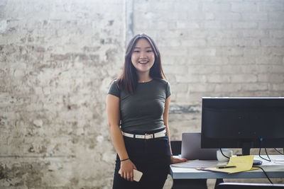 Portrait of smiling female computer programmer standing beside desk at creative office