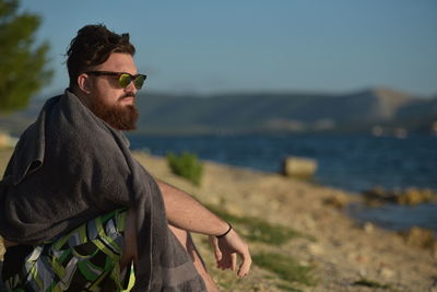 Bearded man sitting at beach against sky