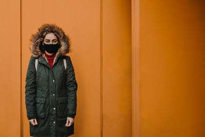 Portrait of young woman wearing mask standing against yellow wall