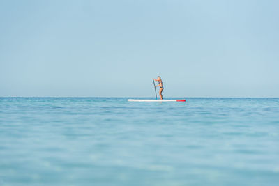 Side view of fit female surfer in bikini standing on paddleboard and rowing while training in sea on sunny day