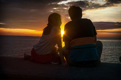 Rear view of couple sitting by sea against sky during sunset