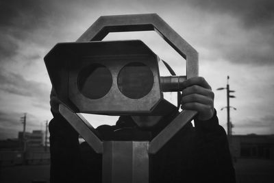 Man using coin-operated binocular against cloudy sky