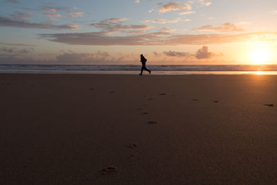 Silhouette man jogging on sand against cloudy sky at beach during sunset