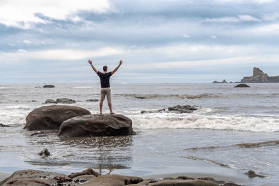 Man with arms raised in triumph of freedom on a pacific coast rocky beach, sand and surf.