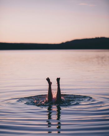 Low section of young woman swimming in lake against sky at sunset