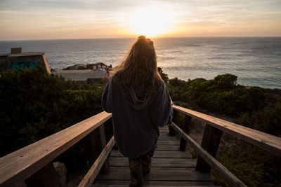 Rear view of woman looking at sea while standing on steps against sky during sunset