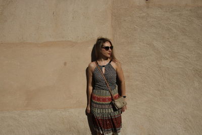 Portrait of young woman wearing sunglasses standing against wall