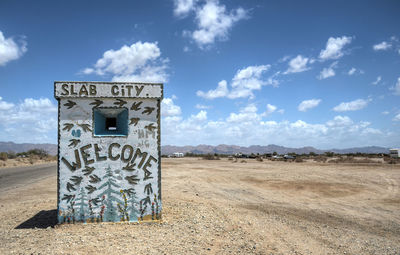 Welcome sign on wall at sandy field against sky