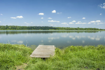 Wooden jetty on the lake, trees on the horizon and white clouds on a blue sky.