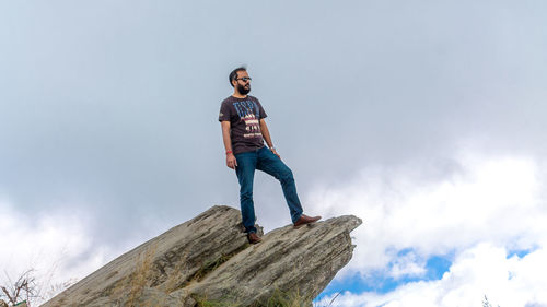 Low angle view of young man standing on rock against sky