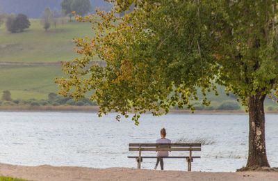 Young woman sitting on a bench under a tree in front of a lake in the evening in autumn