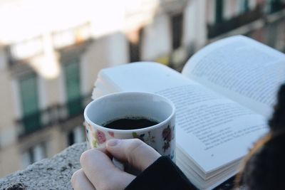 Cropped hand of woman holding coffee cup with book on railing