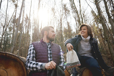 Low angle view of mature male and teenager sitting on log in forest