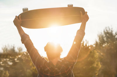 Rear view of man with arms raised holding skateboard during sunset
