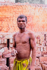Portrait of shirtless man standing against brick wall