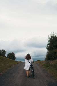 Rear view of woman walking with bicycle on road against sky