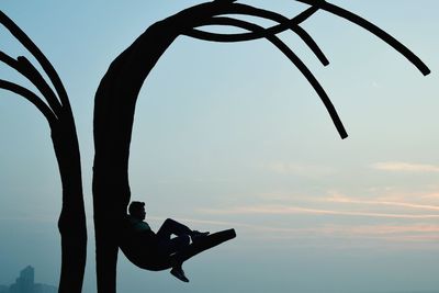 Silhouette man sitting on artificial tree against sky during sunset