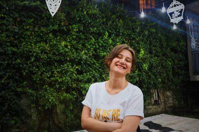 Portrait of smiling young woman standing against plants in city