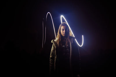 Portrait of young woman standing against light painting at night
