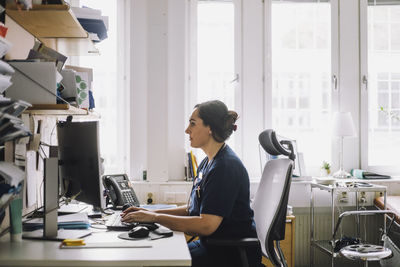 Side view of focused female healthcare worker using computer while sitting on desk in clinic