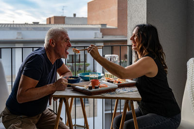 Beautiful middle aged couple eating chinese take away food sitting at a laid table in a city balcony