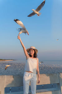 Beautiful woman wearing hat feeding seagull while standing against sky