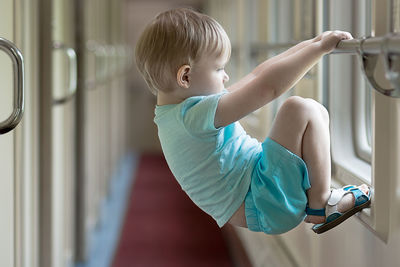 A bored and sad blond boy toddler hangs on the crossbar of a train window. 