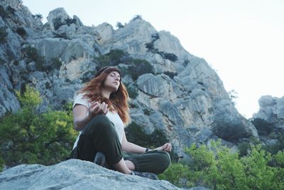 Low angle view of young woman meditating while sitting on rock