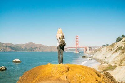 Full length rear view of woman standing on rock at sea shore by golden gate bridge against clear blue sky