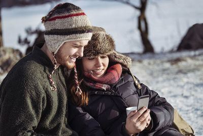 Smiling couple using smart phone while relaxing on snow covered field