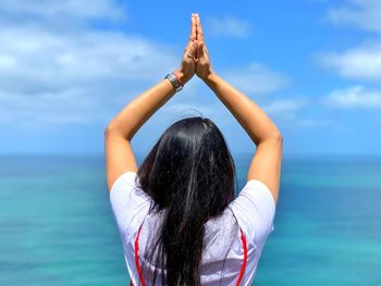 Rear view of woman with arms raised and hands clasped standing by sea against sky