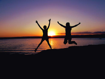 Silhouette women with arms raised jumping over beach against sky during sunset