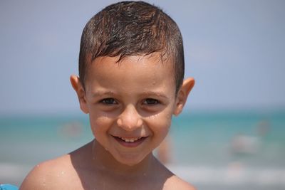 Close-up portrait of smiling shirtless boy standing against sea during sunny day