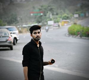 Portrait of young man holding camera lens while standing on road