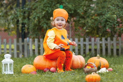 Full length of cute girl wearing costume sitting by pumpkin at park